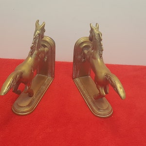 May include: A pair of gold-colored metal bookends, each featuring a sculpted horse in a rearing position. The horses are attached to a flat, oval-shaped base with a vertical support plate behind them, showcasing a metallic finish and detailed manes.