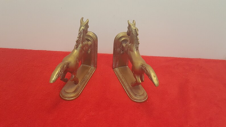 May include: A pair of gold-colored metal bookends, each featuring a sculpted horse in a rearing position. The horses are attached to a flat, oval-shaped base with a vertical support plate behind them, showcasing a metallic finish and detailed manes.