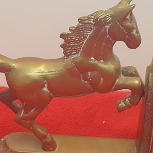May include: A pair of brass-colored metal bookends featuring a horse in a rearing position. Each horse is attached to a curved, solid base with a vertical support plate. The metallic finish shows signs of wear and texture, with a distinct horse silhouette.