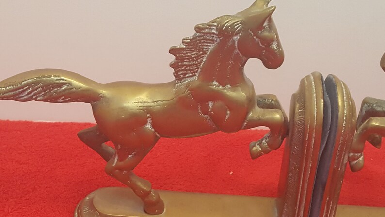 May include: A pair of brass-colored metal bookends featuring a horse in a rearing position. Each horse is attached to a curved, solid base with a vertical support plate. The metallic finish shows signs of wear and texture, with a distinct horse silhouette.