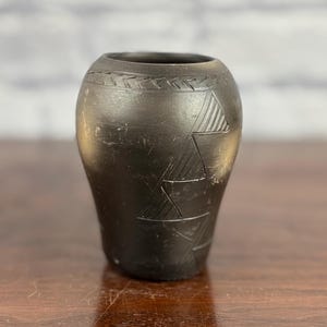 May include: A small, dark-colored ceramic vase with etched geometric patterns. The vase has a rounded body and a slightly flared opening. The surface appears to be matte, and the vase is sitting on a wooden surface.