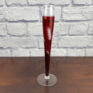 May include: A tall, slender glass with a red liquid inside, possibly wine or a cocktail. The glass has a clear base and a decorative etched feather design. The background is a white brick wall.