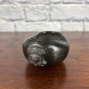 May include: A small, polished black ceramic bowl with a rounded shape and a smooth, glossy finish. The exterior features a molded relief of a stylized human face with a detailed headdress centered on the front side of the vessel.