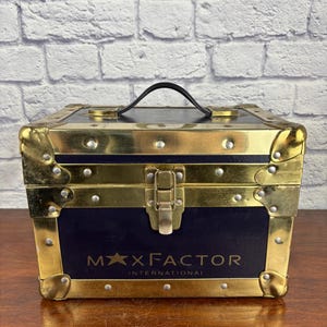 Op de afbeelding: Een marineblauwe en gouden Max Factor International make-upkoffer. De koffer heeft gouden hoekbeschermers, een gouden sluiting en een zwarte handgreep. De tekst "MAX FACTOR INTERNATIONAL" staat op de voorkant.