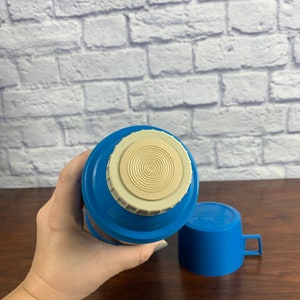 May include: Blue and white thermos with a lid and a matching cup. The thermos has a white lid with a circular design and the text "Thermos" on it.