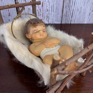 以下が含まれることがあります： 素朴な木製のゆりかごの中にいる赤ちゃんの置物。赤ちゃんは白いおむつをしており、光輪があります。ゆりかごは白い毛皮で裏打ちされています。背景は明るい色の木製の表面です。