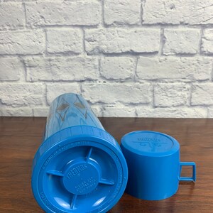 May include: A blue Thermos vacuum bottle with a silver lid and a blue cup. The bottle has the text "A Brand of Vacuum Bottle" and "Thermos" printed on it. The cup has the text "Thermos" printed on it.