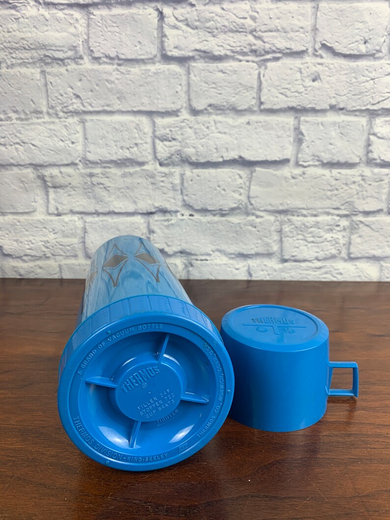 May include: A blue Thermos vacuum bottle with a silver lid and a blue cup. The bottle has the text "A Brand of Vacuum Bottle" and "Thermos" printed on it. The cup has the text "Thermos" printed on it.