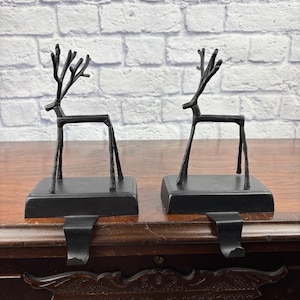 May include: A pair of black metal stocking holders, each featuring a stylised reindeer silhouette standing on a rectangular base with a curved hook extending from the front to support a stocking or other hanging item.