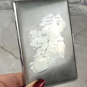 May include: Silver cigarette case with a textured diamond pattern. The case features an etched map of Ireland, with city names like Dublin and Belfast. The case is rectangular and has a hinged opening.