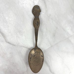 May include: A tarnished brass spoon featuring an elongated handle shaped like a human figure with a profile face at the top. The bowl of the spoon displays etched markings, including a sunburst pattern at the base and worn, dark surface textures.