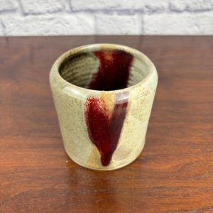 Puede incluir: Una taza de cerámica hecha a mano con un exterior beige y un diseño en forma de corazón rojo oscuro. El interior es oscuro. La taza es cilíndrica con un borde ligeramente ensanchado. La cerámica tiene un aspecto rústico y texturizado.