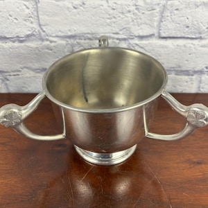 Puede incluir: Una taza de metal plateado con dos asas ornamentadas y un asa pequeña en la parte superior. La taza tiene una abertura ancha y se asienta sobre una base pequeña y redonda. Las asas tienen un diseño floral decorativo.