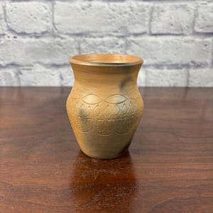 May include: A small, handcrafted pottery vase with a light brown exterior and a darker rim. The vase features a decorative pattern of repeating shapes around the middle. The vase is displayed on a dark wooden surface.