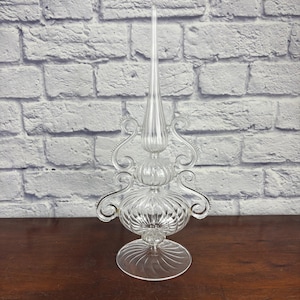 May include: A clear glass Christmas tree topper with a long, pointed spire. The decorative piece features a bulbous base with swirling details and ornate, curved accents. The topper is displayed on a dark wooden surface against a white brick wall.