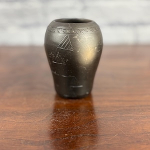 Puede incluir: Un pequeño jarrón gris oscuro con diseños geométricos grabados. El jarrón tiene un cuerpo redondeado y una abertura ancha. Los diseños grabados incluyen un triángulo y otras formas. El jarrón está sobre una superficie de madera.