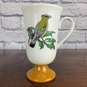 May include: White ceramic pedestal mug with a yellow and green bird illustration on the front. The bird is perched on a leafy branch with red berries. The base of the mug is a solid orange color.