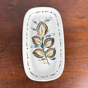 May include: White ceramic serving dish with a hand-painted botanical design. The rectangular dish features a central motif of stylized leaves in brown, yellow, and blue, with a border of green leaves and black dots. A decorative piece for serving.