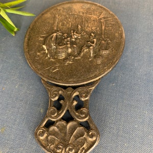 May include: A silver-toned hand mirror with an ornate handle. The mirror features an embossed scene of people gathered around a barrel.