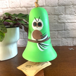 May include: Green ceramic owl bell with a brown and white painted owl design. The bell is hanging from a brown leather cord and is sitting on a brown wooden surface. The bell is labeled "Tesia Southwest Originals".