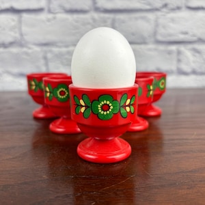 May include: A white egg sits in a red egg cup with a floral design. The egg cup is part of a set of four, each with a similar green and yellow floral pattern. The egg cups are on a dark wooden surface.