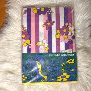 May include: A package of bed linen featuring vertical stripes in white, light pink, and dark purple, accented with floral patterns in yellow, pink, and purple. The packaging includes the text Modische Bettwäsche and a graphic of a person's head.