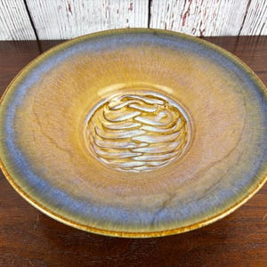 May include: A ceramic bowl with a textured, swirling design in the center. The bowl has a gradient of colors, with a golden-brown interior, a blue-grey rim, and a light brown exterior. The bowl is round and sits on a wooden surface.
