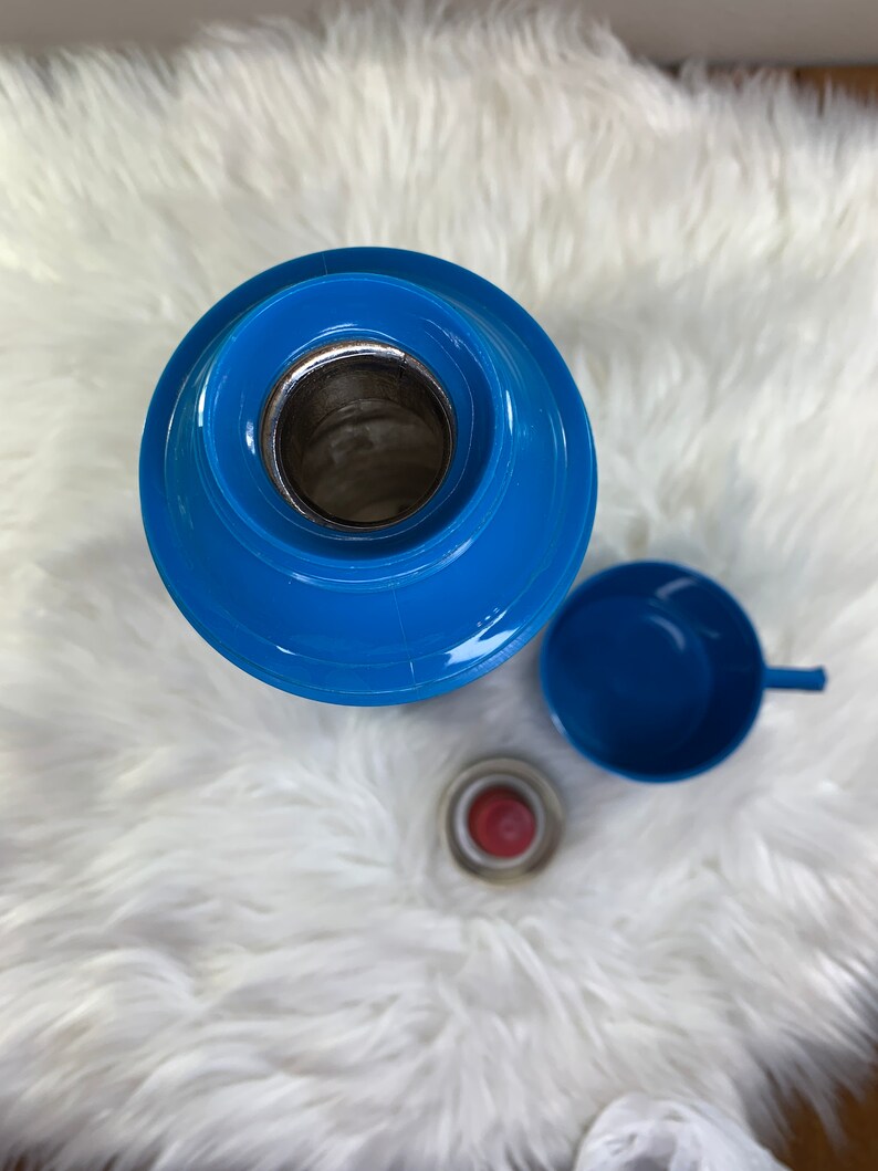 May include: A blue thermos with a blue lid and a blue cup. The thermos is on a white fluffy surface.