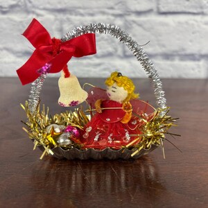 Pode incluir: Enfeite de Natal vintage com um pequeno anjo em um vestido vermelho com cabelo dourado, sentado em uma cesta. A cesta é decorada com fios dourados e pequenos enfeites. Um laço vermelho e um sino penduram-se na alça prateada.