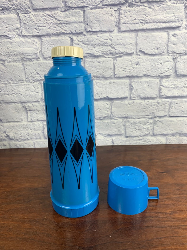 May include: Blue and black thermos with a diamond pattern and a matching blue cup. The thermos has a white lid and the cup has a handle.