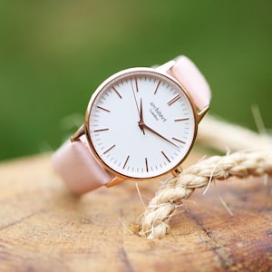 May include: A rose gold wristwatch with a white face and pink leather band. The watch face has roman numeral markings and the text "architect London".