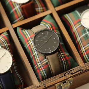 May include: A black watch with a gray leather band in a wooden box with red and green plaid fabric lining. The watch face has white hands and the text "architect London" printed on it.