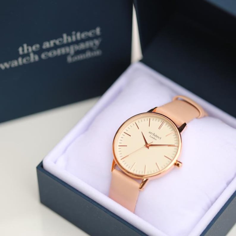 May include: A rose gold watch with a white face and rose gold hands. The watch has a pink leather band and is in a blue box with the text "the architect watch company london".
