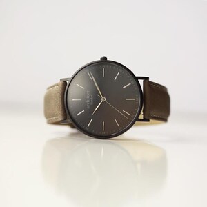 May include: A black wristwatch with a brown leather band. The watch face is black with white markings and the word "architect London" printed on it.