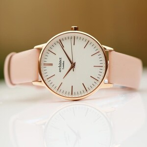May include: A rose gold watch with a white face and a pink leather band. The watch face has the words "architect London" printed on it.