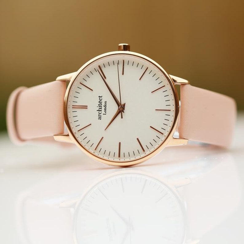 May include: A rose gold watch with a white face and a pink leather band. The watch face has the words "architect London" printed on it.