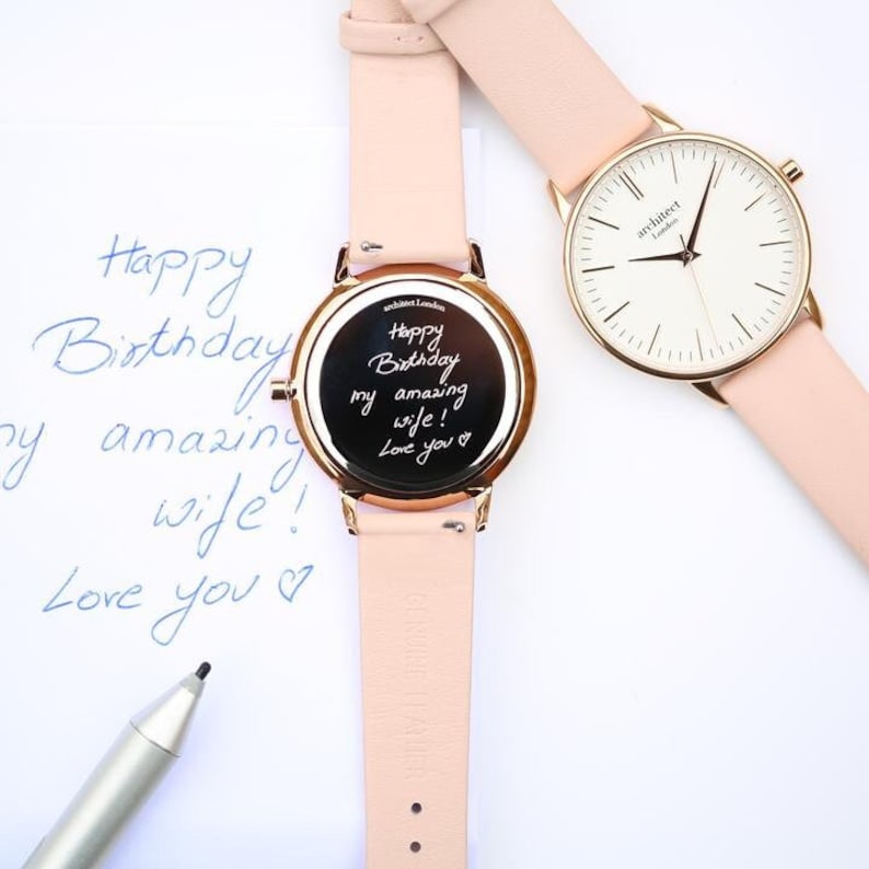 May include: A pink leather wristwatch with a gold-tone case. The back of the watch is engraved with the message "Happy Birthday my amazing wife! Love you".