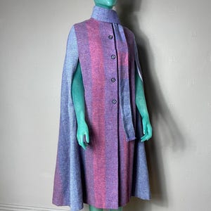 May include: A vintage cape in shades of purple and pink, displayed on a mannequin. The cape features a vertical striped pattern and a matching scarf. It has a button-up front and a cape-style design with arm openings.