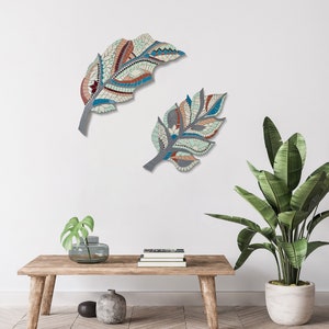 May include: Two mosaic feather wall art pieces. The feathers are in shades of blue, orange, and cream, with a gray stem. The mosaic design is intricate and detailed, creating a unique decorative element for any room. The feathers are mounted on a white wall.