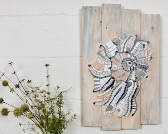 Può includere: Un'opera d'arte murale a mosaico con un design a conchiglia in tonalità di bianco, grigio e rosa. La conchiglia è realizzata con piccole piastrelle ed è montata su uno sfondo di assi di legno.