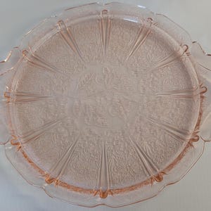 May include: A round, light pink pressed glass serving tray featuring two integrated handles on opposite sides. The surface displays a detailed embossed floral pattern with radiating panels and a scalloped edge, creating a textured appearance.