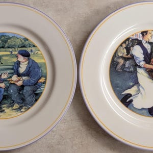 以下が含まれることがあります： 白い背景、黄色のトリム、青いアクセントの2つの装飾プレート。1つのプレートには、公園で会話するベレー帽の2人の男性が描かれています。もう1つは、パンのバスケットを運ぶウェイターを示しています。