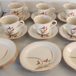 May include: A vintage cream-colored teacup set with matching saucers and plates. Each piece features a gold rim and a stylized floral design in shades of brown and gold. The set includes six cups, saucers, and plates.