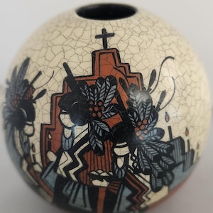 Puede incluir: Un jarrón esférico de cerámica con acabado craquelado. El jarrón presenta un diseño con una cruz, elementos florales y formas geométricas en negro, naranja y azul sobre un fondo crema. El jarrón tiene un interior negro.