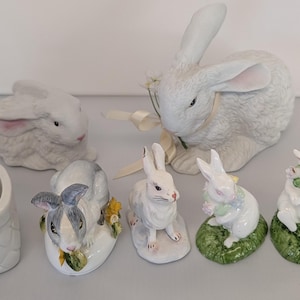 May include: A collection of ceramic Easter bunny figurines in various poses and sizes. The bunnies are primarily white with pink accents, some with floral decorations and baskets. A white basket with a bunny attached is also present.