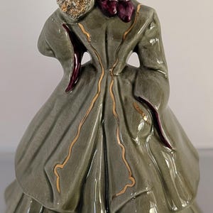 May include: A glazed ceramic figurine of a person wearing a long, olive-green coat with gold trim, a deep purple bonnet, and a dark purple scarf. The figure holds a textured, gold-accented bouquet near their face, featuring painted facial details.