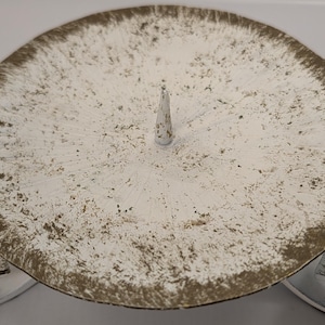 May include: A distressed white metal candle holder with a circular base and a central spike. The rim and the two curved feet have a brown, aged finish. The candle holder is designed to hold a single candle.