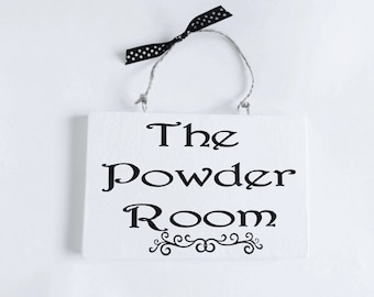 May include: A white rectangular sign with the words "The Powder Room" in black script. The sign is decorated with a black and white polka dot ribbon and a decorative flourish at the bottom.