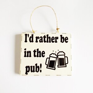 Peut inclure: Une pancarte en bois blanc avec du texte noir qui dit "J'aimerais mieux être au pub !" et un graphique de deux chopes de bière qui trinquent. La pancarte a une boucle en corde pour l'accrocher.