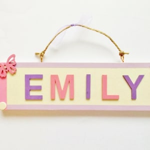 Puede incluir: Un letrero de madera personalizado con el nombre "EMILY" en letras moradas y rosas. El letrero está decorado con mariposas rosas y moradas y tiene una cuerda de yute para colgar. El fondo es de color amarillo claro.